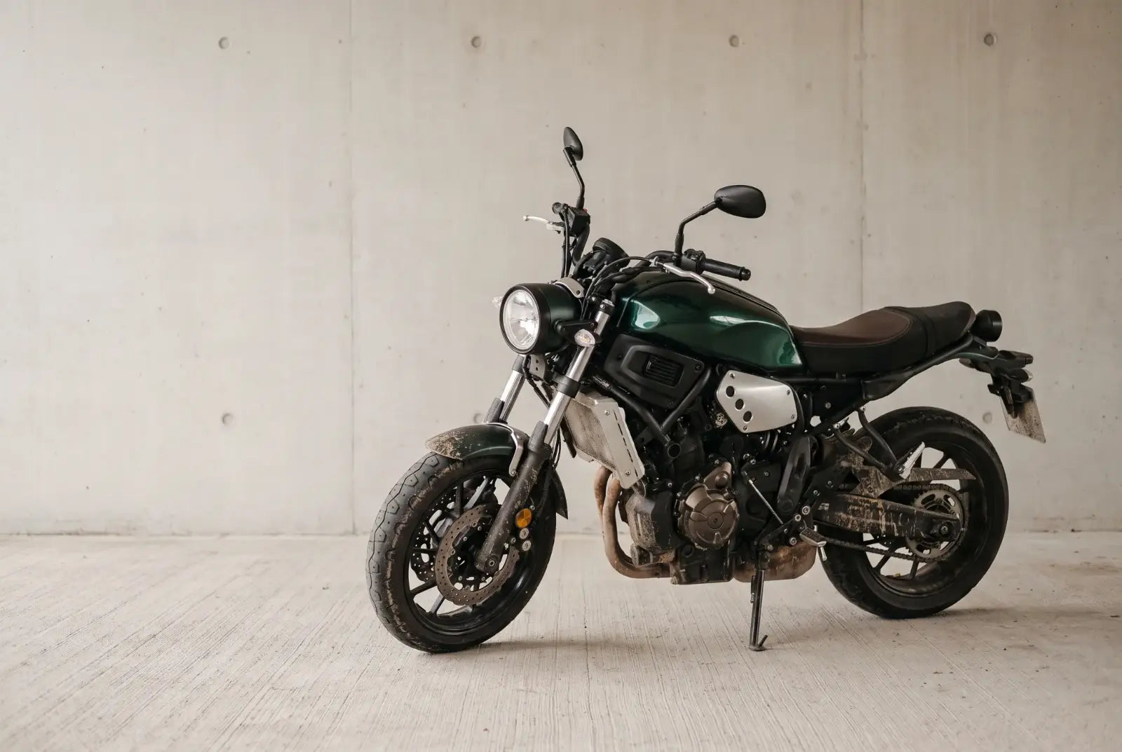 Moto verde de estilo clássico estacionada diante de uma parede de concreto claro, com poeira e respingos de lama nas rodas e no motor